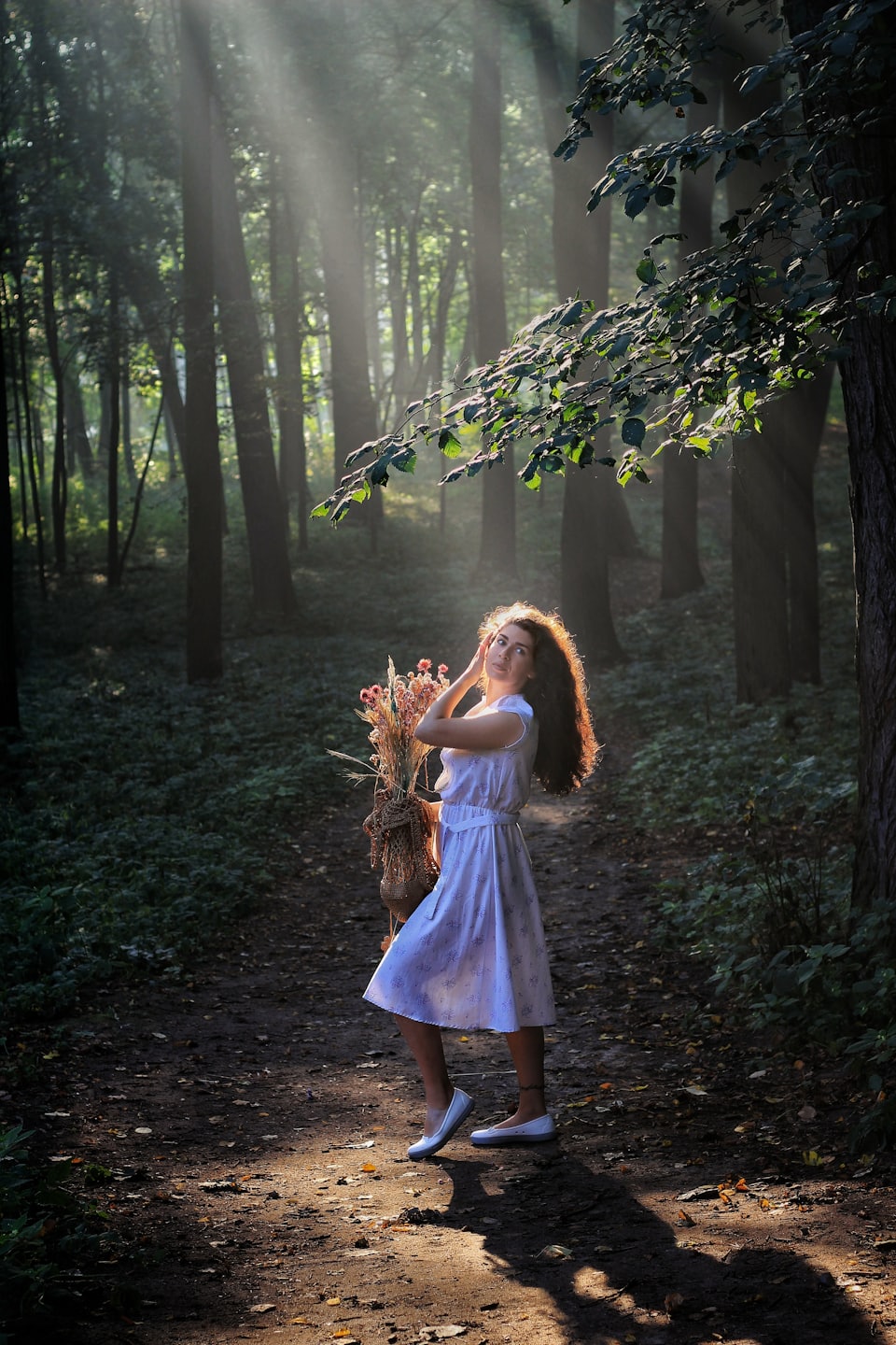 Woman in a white dress holds dried flowers on a forest path.